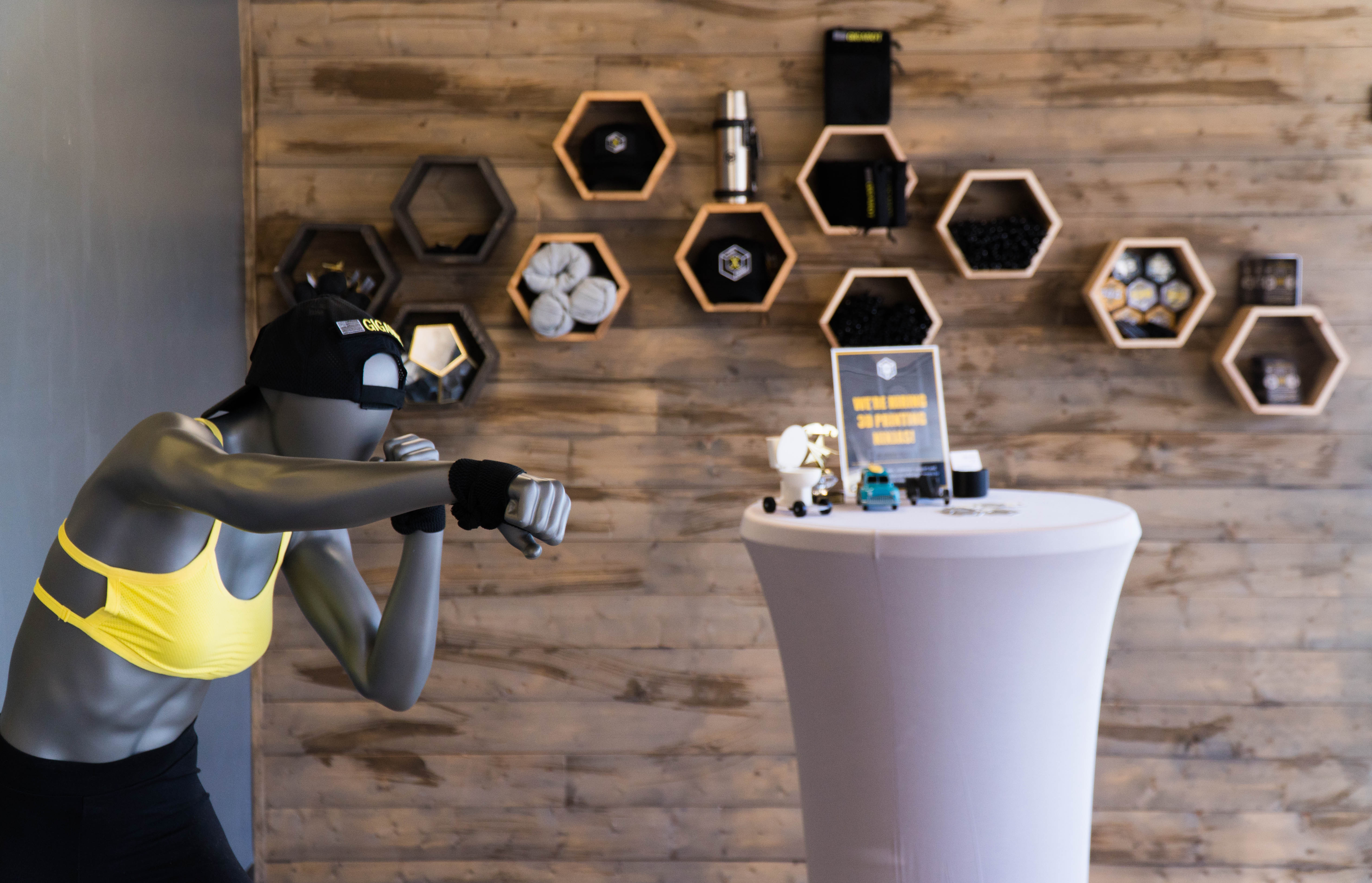 A silver mannequin of a boxing lady wearing a black hat and yellow tank top stands in front of a rustic wooden slat wall holding re:3D merchandise.