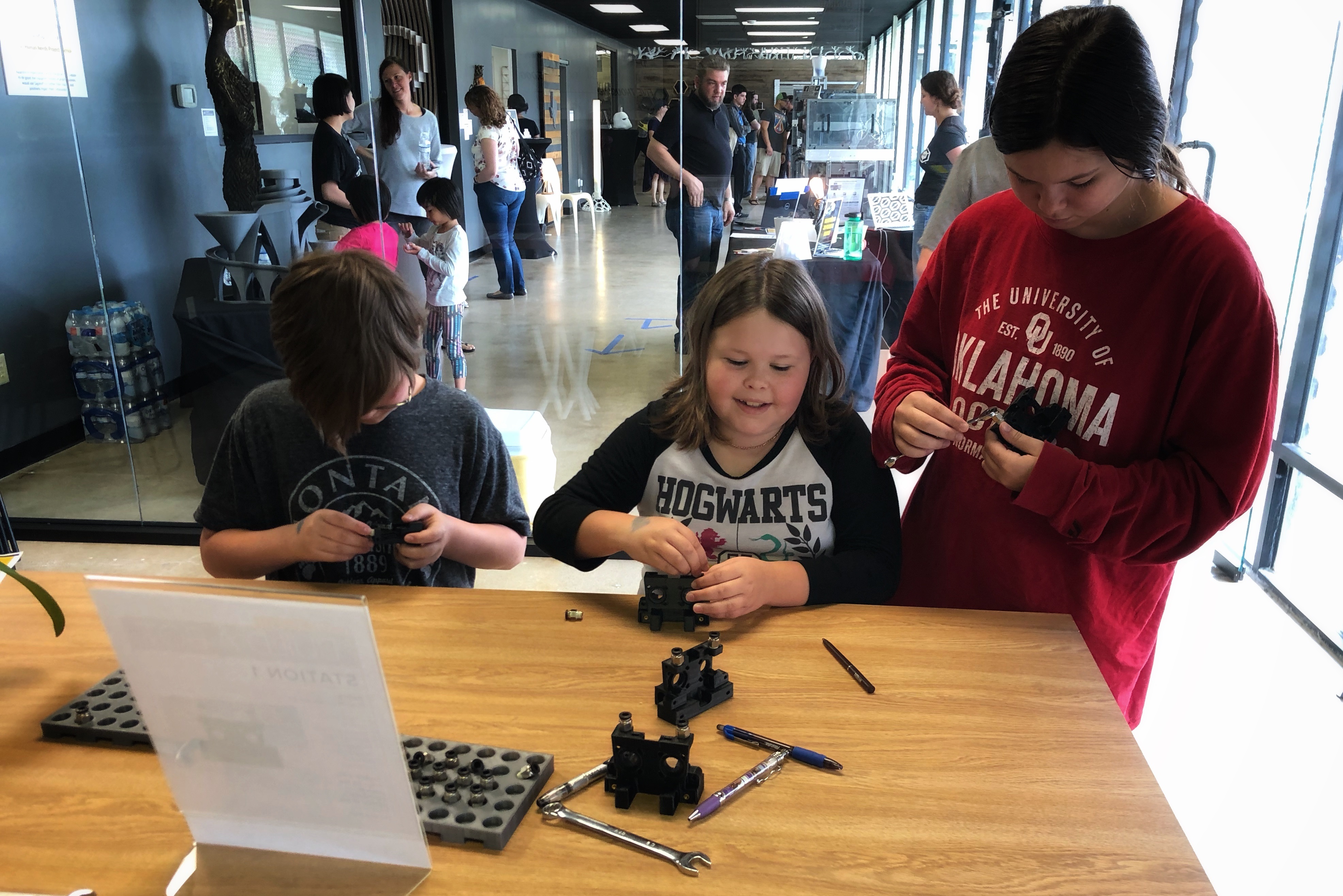 Three kids sit at a table with tools and build part of a 3D printer. A crowd of people is in the background.