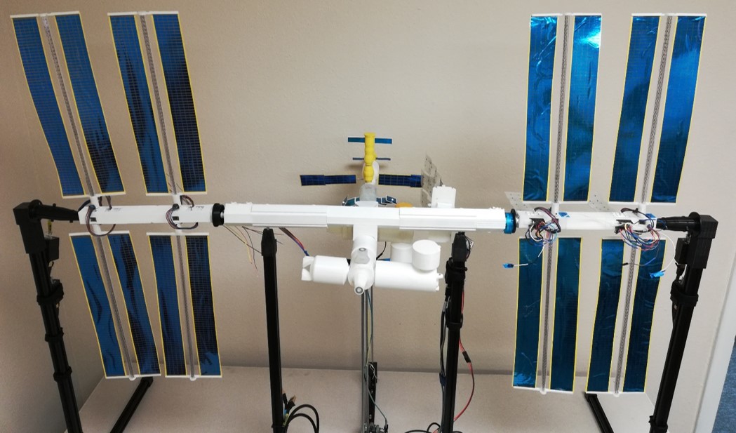 ISS Mimic, a 100th scale model of ISS, sits on a table. You can see 3d printed tubes, wires connecting to motors and large, foil covered solar arrays.