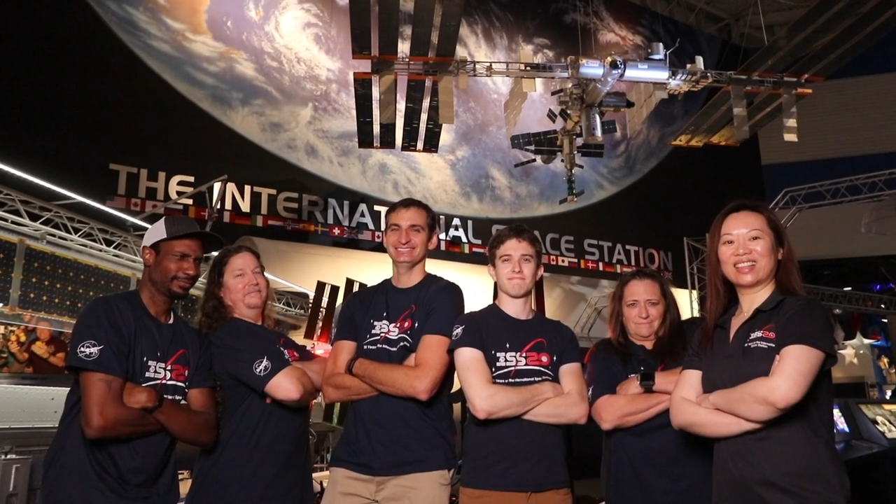 Six people, four men and two women stand in front of a display of the earth with the ISS floating above their heads. All of them are wearing tshirts that read "ISS20"